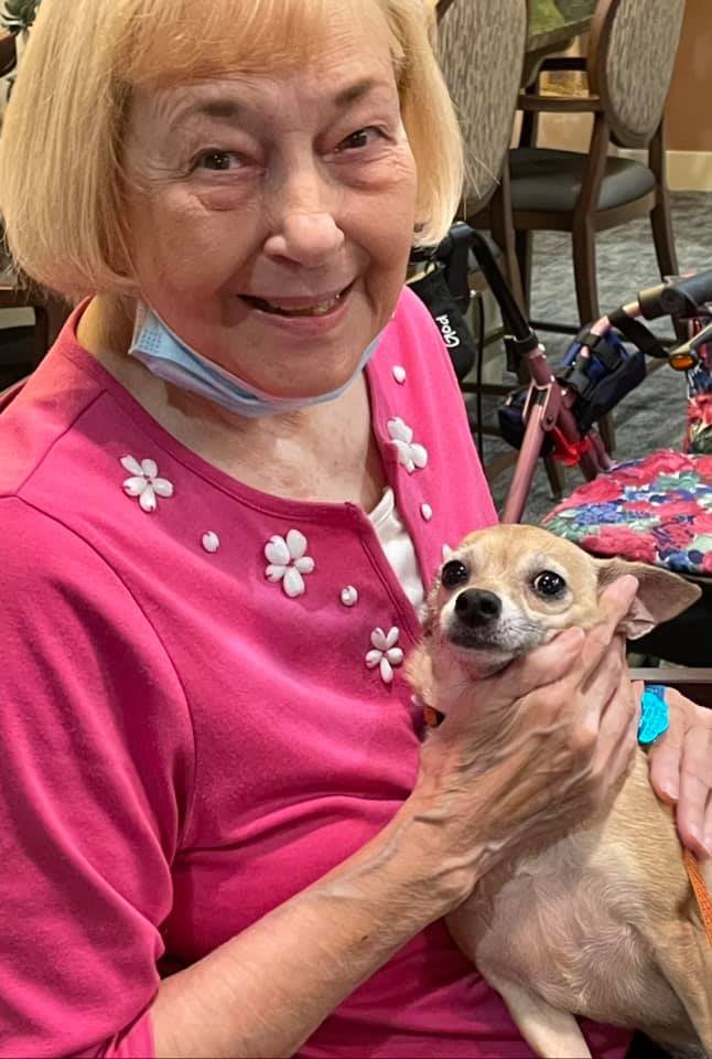 An elderly woman smiling while holding a small dog indoors.