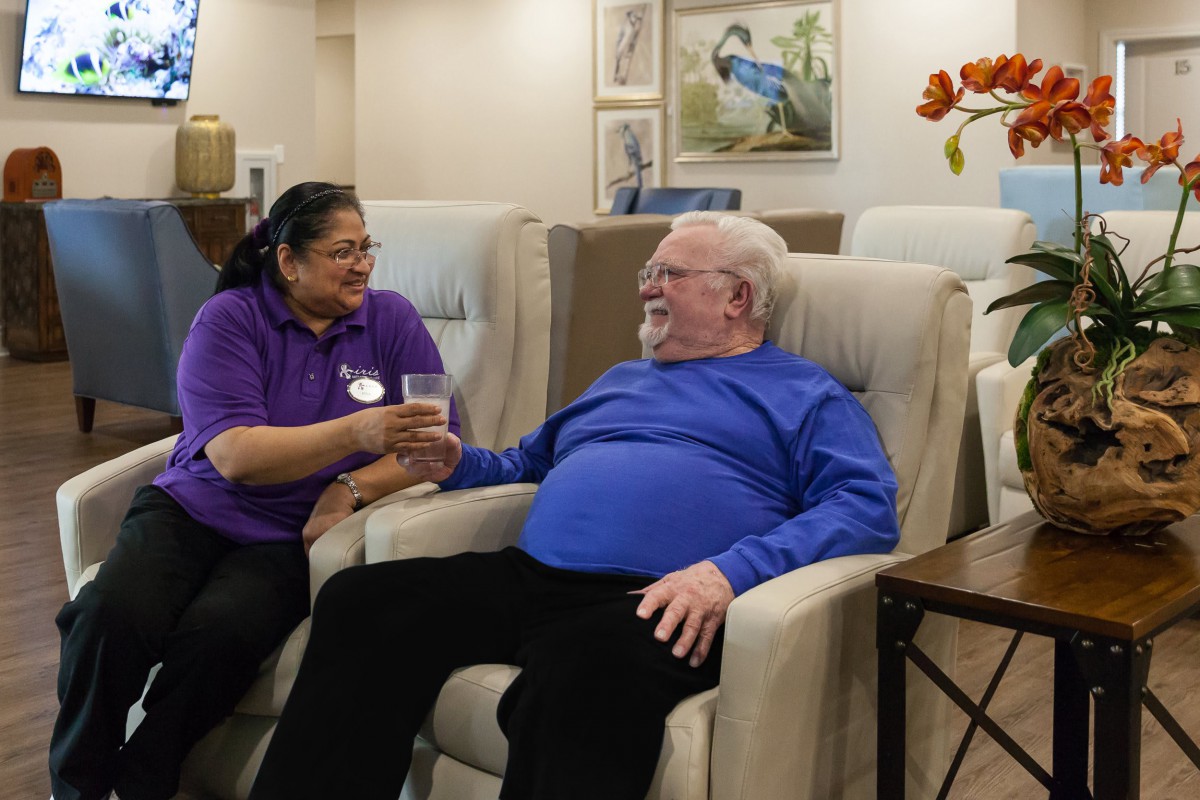 An elderly man sits comfortably in a chair while a caregiver offers him a drink, showcasing a moment of friendly interaction in a care facility.