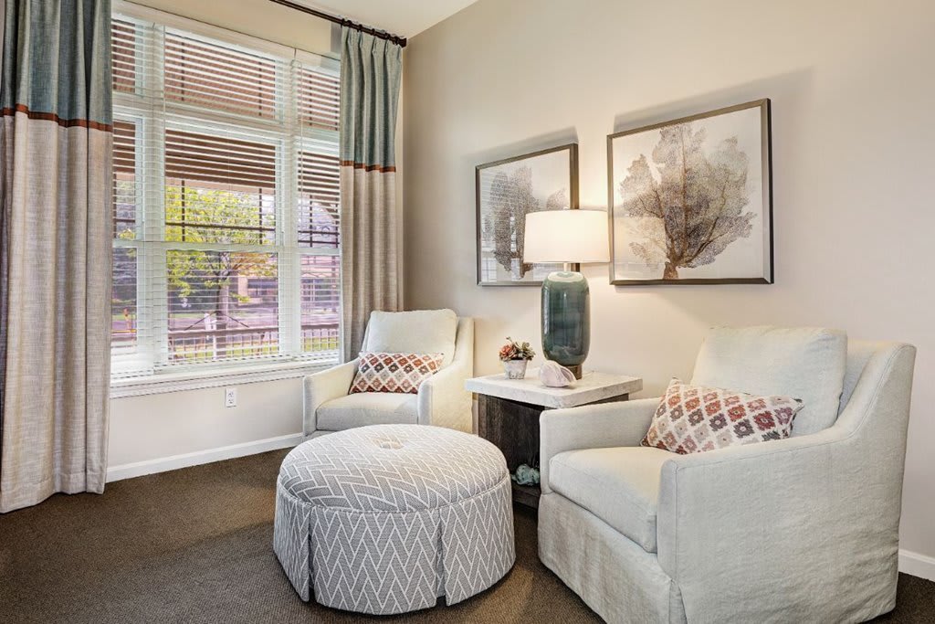A cozy living room corner featuring two armchairs, a round ottoman, a table lamp, and two pieces of artwork, with abundant natural light coming through the window.