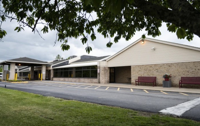 A view of a school building with a parking area and a bench nearby.