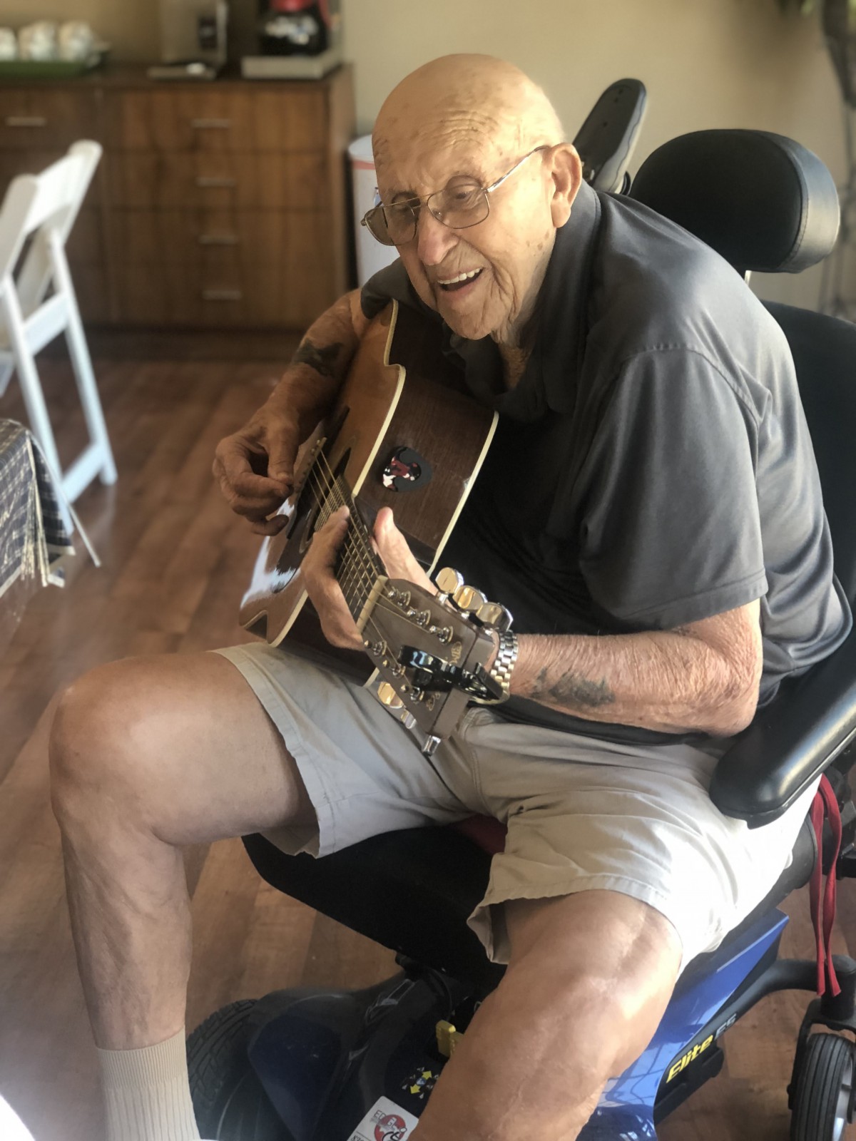 A smiling man sits indoors playing a guitar.