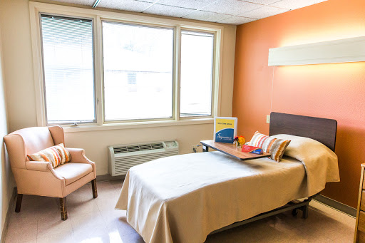 A bright and comfortable hospital room featuring a bed, a chair, and natural light streaming through the windows.