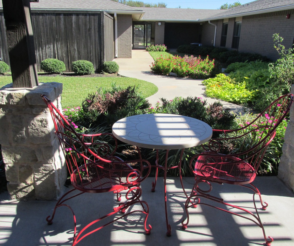 A sunny patio with red chairs and a round table overlooking a landscaped garden.