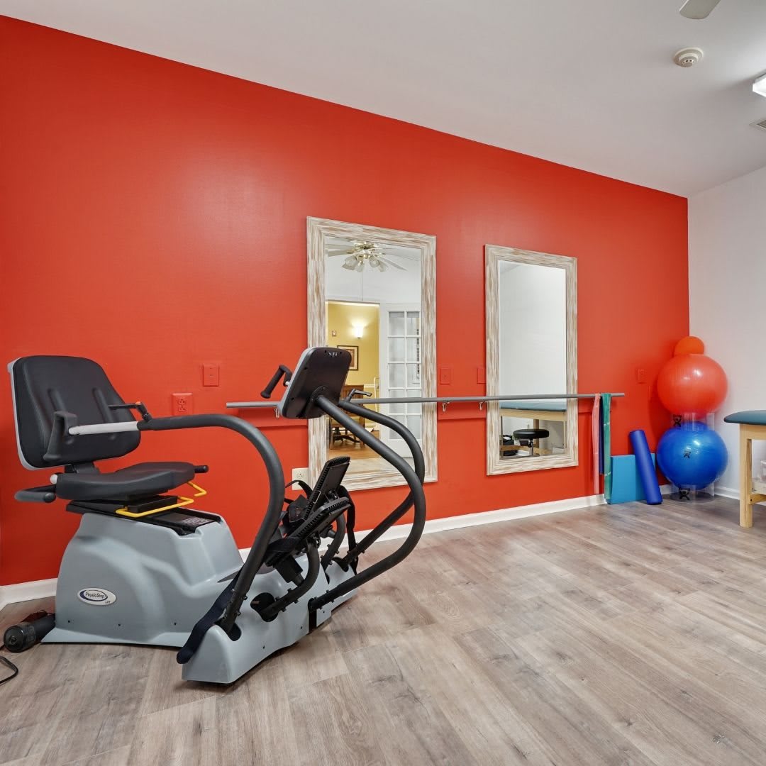 A rehabilitation room featuring a recumbent exercise bike, mirrors, and exercise balls against a red wall.