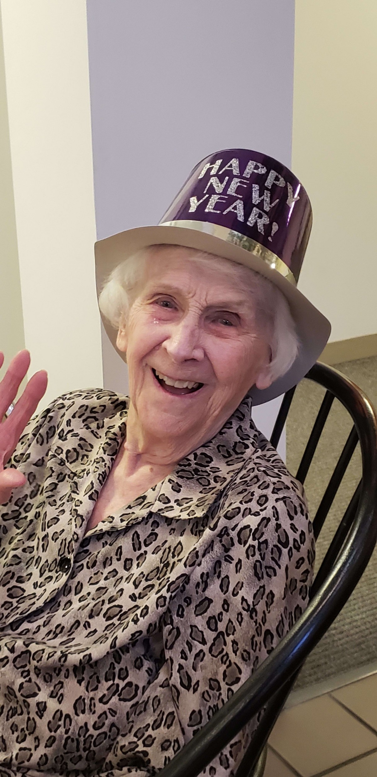An elderly woman smiling and waving while wearing a party hat and leopard print top.