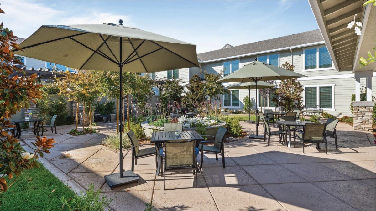 A sunny outdoor patio area with tables, chairs, and umbrellas surrounded by a landscaped garden.