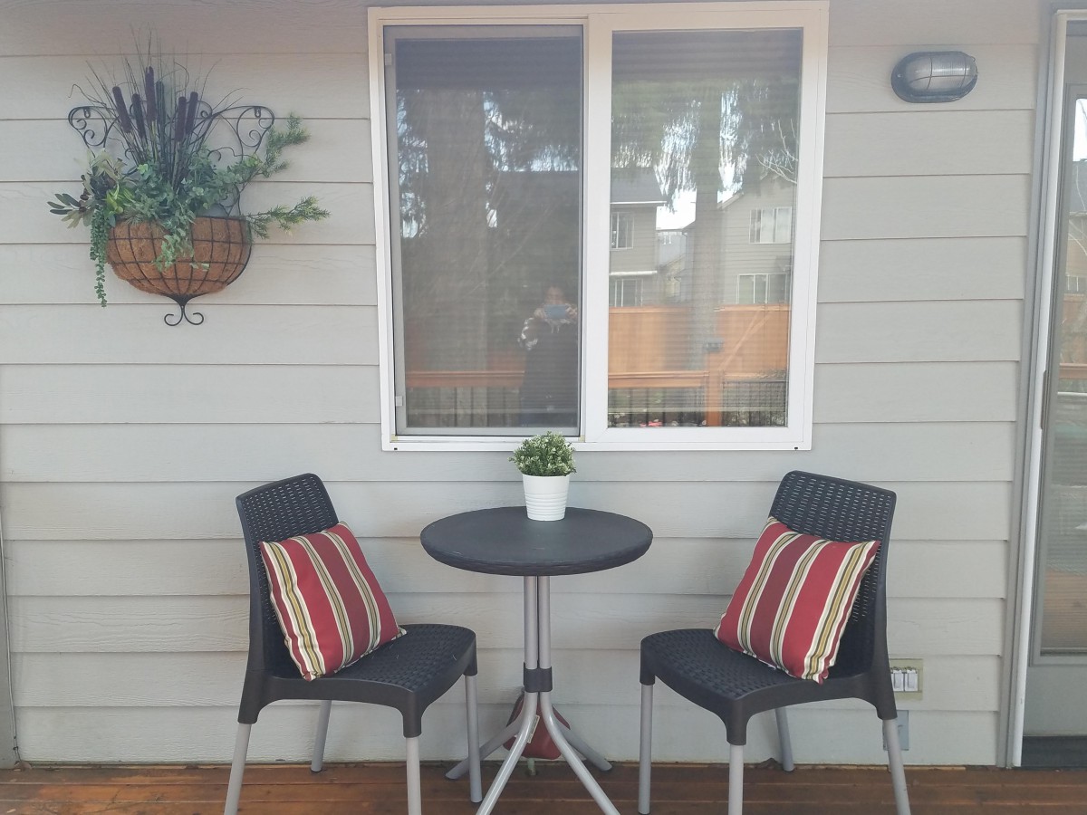 A cozy outdoor patio setup with a small table and two chairs adorned with striped cushions, set against a house wall with a window and hanging plants.