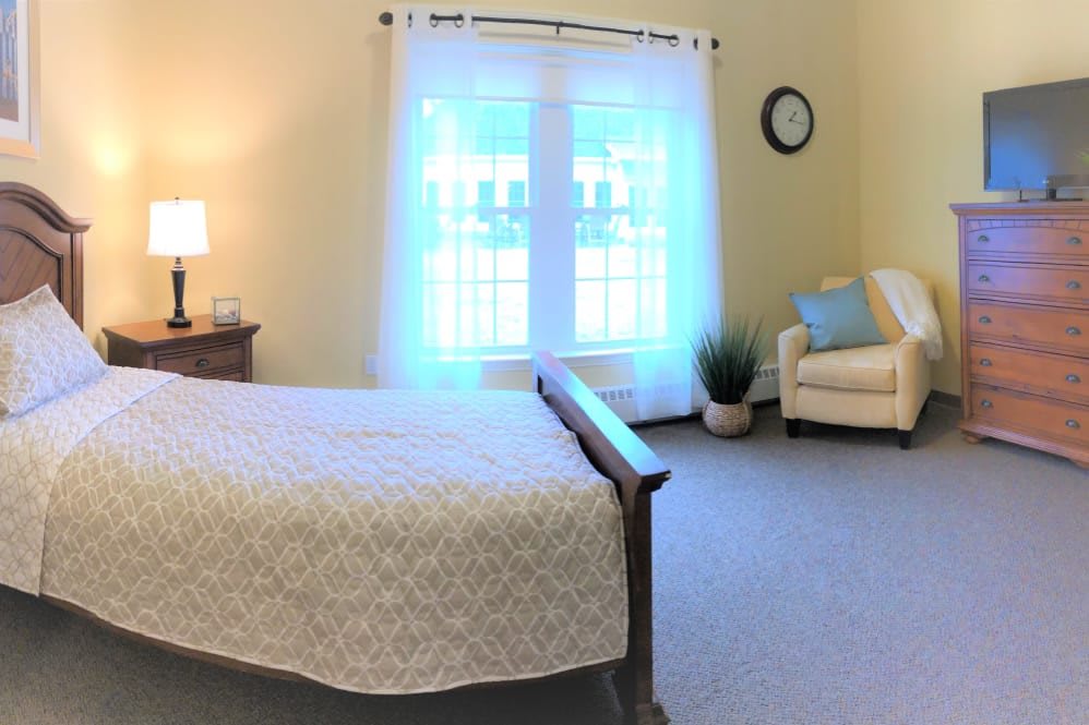 A cozy bedroom featuring a bed with a light-colored quilt, an armchair, and large windows with sheer curtains.