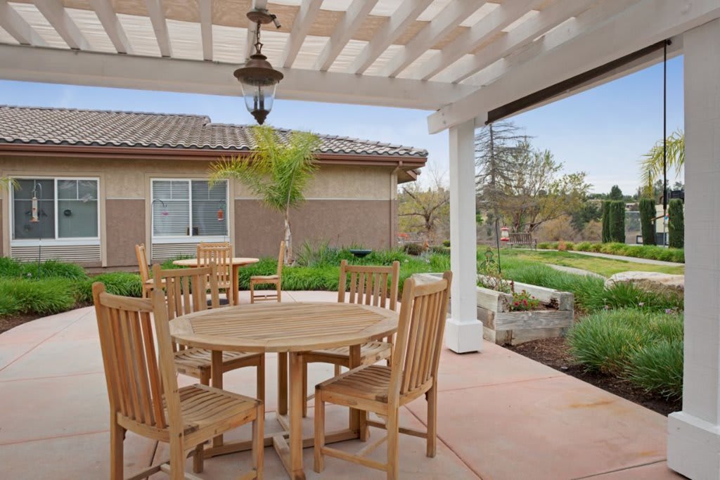 A cozy backyard patio with a round table and chairs under a pergola, surrounded by garden landscaping.