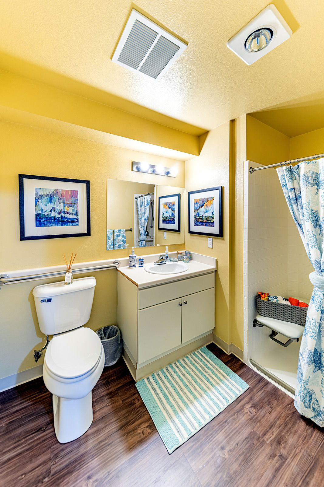 A small, brightly lit bathroom featuring a toilet, sink with storage, and a shower with a patterned curtain.