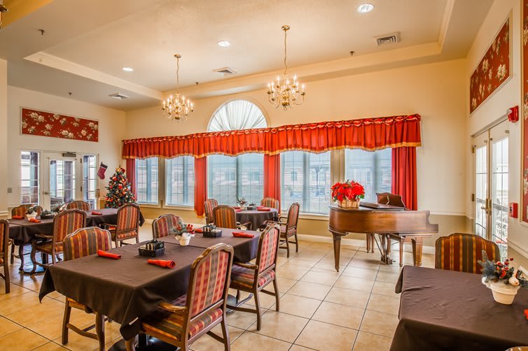 A well-lit dining area with multiple tables and chairs, featuring large windows with red curtains and elegant chandeliers.