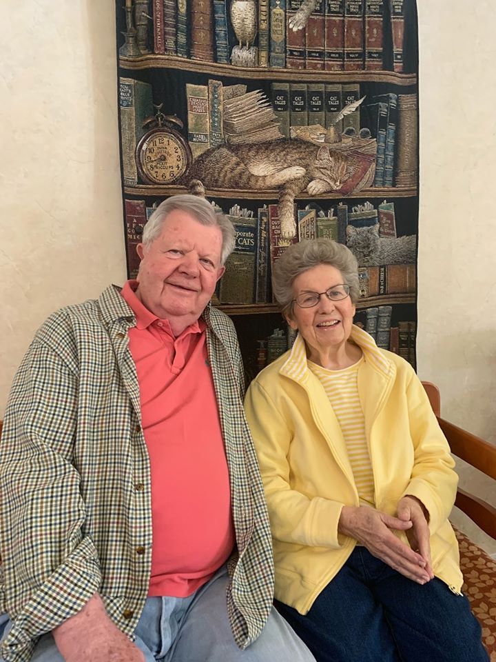 Elderly couple sitting together indoors with a bookshelf in the background.
