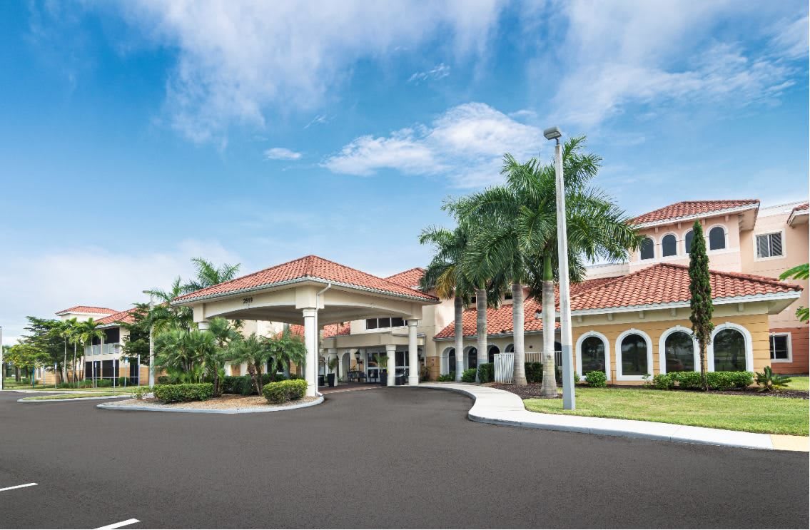 A grand building entrance with palm trees lining the driveway under a clear blue sky.