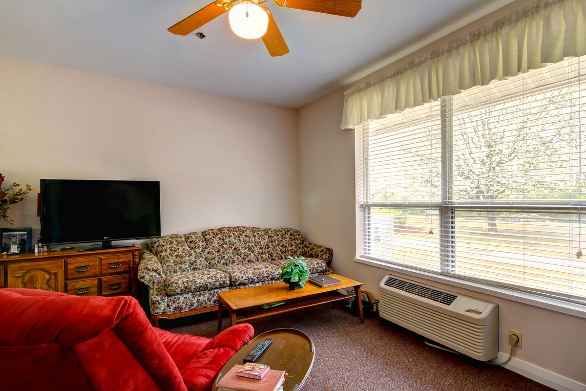 A cozy living room with a patterned sofa, red armchair, and coffee table facing a television. The room includes a ceiling fan, large window with curtains, and an air conditioning unit.