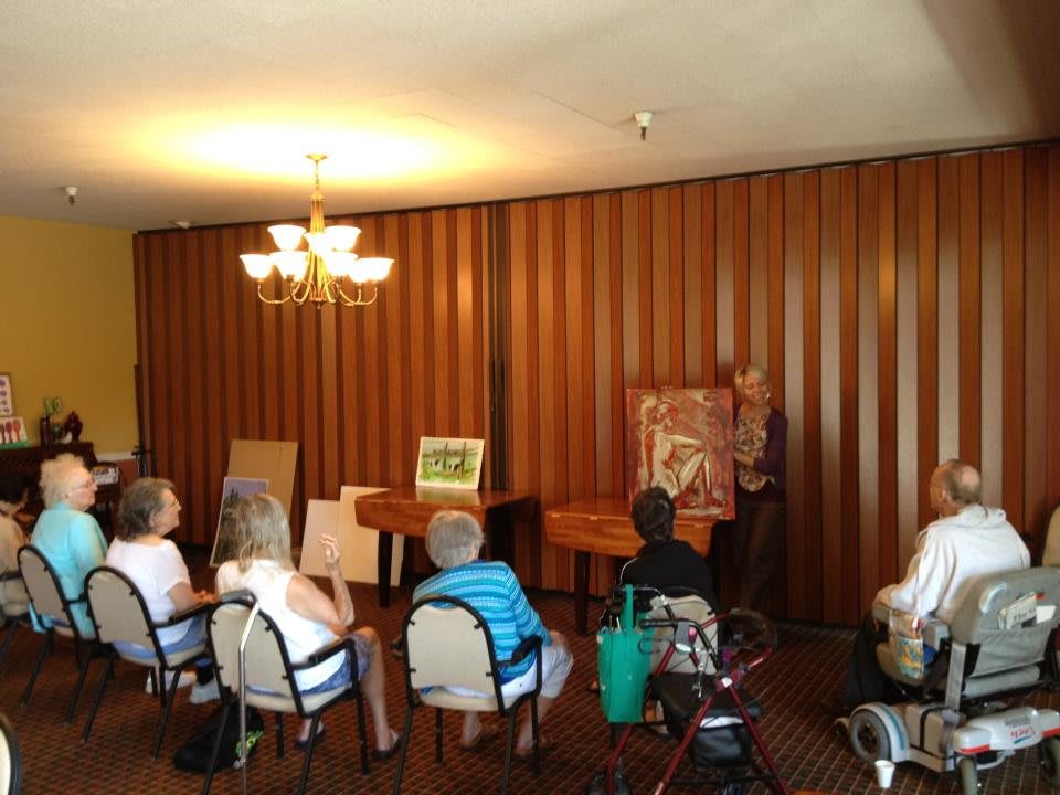 A small group of people seated inside, viewing an art exhibition with displayed paintings.
