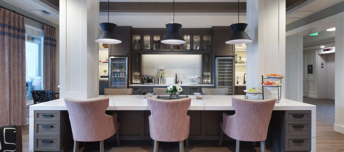 A stylish modern kitchen featuring a central island with pink chairs, accented by pendant lights and dark cabinets.