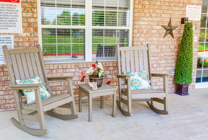 A cozy patio setup with two wooden rocking chairs, a small table, and decorative pillows against a brick wall.