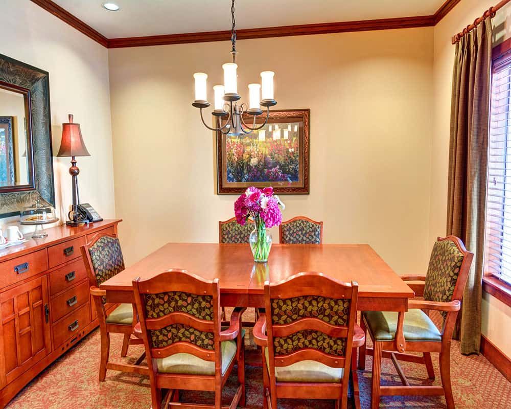 A warmly lit dining room featuring a wooden table with six chairs, a chandelier overhead, and a decorative painting on the wall.