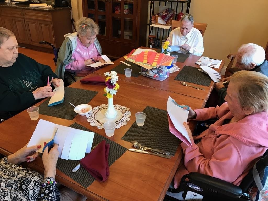A group of elderly individuals engaged in a writing activity around a table indoors.
