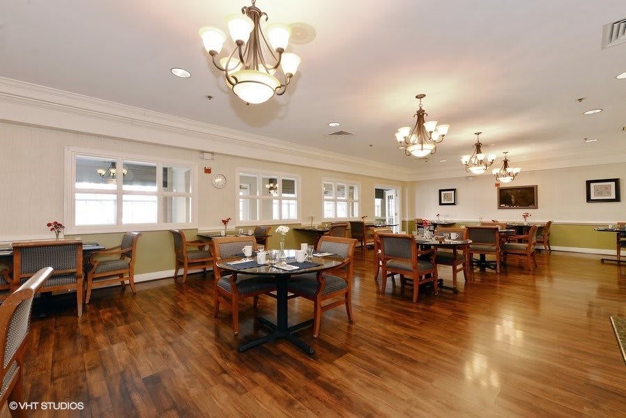A spacious dining room with wooden floors, furnished with multiple tables and chairs, elegantly lit by chandeliers.