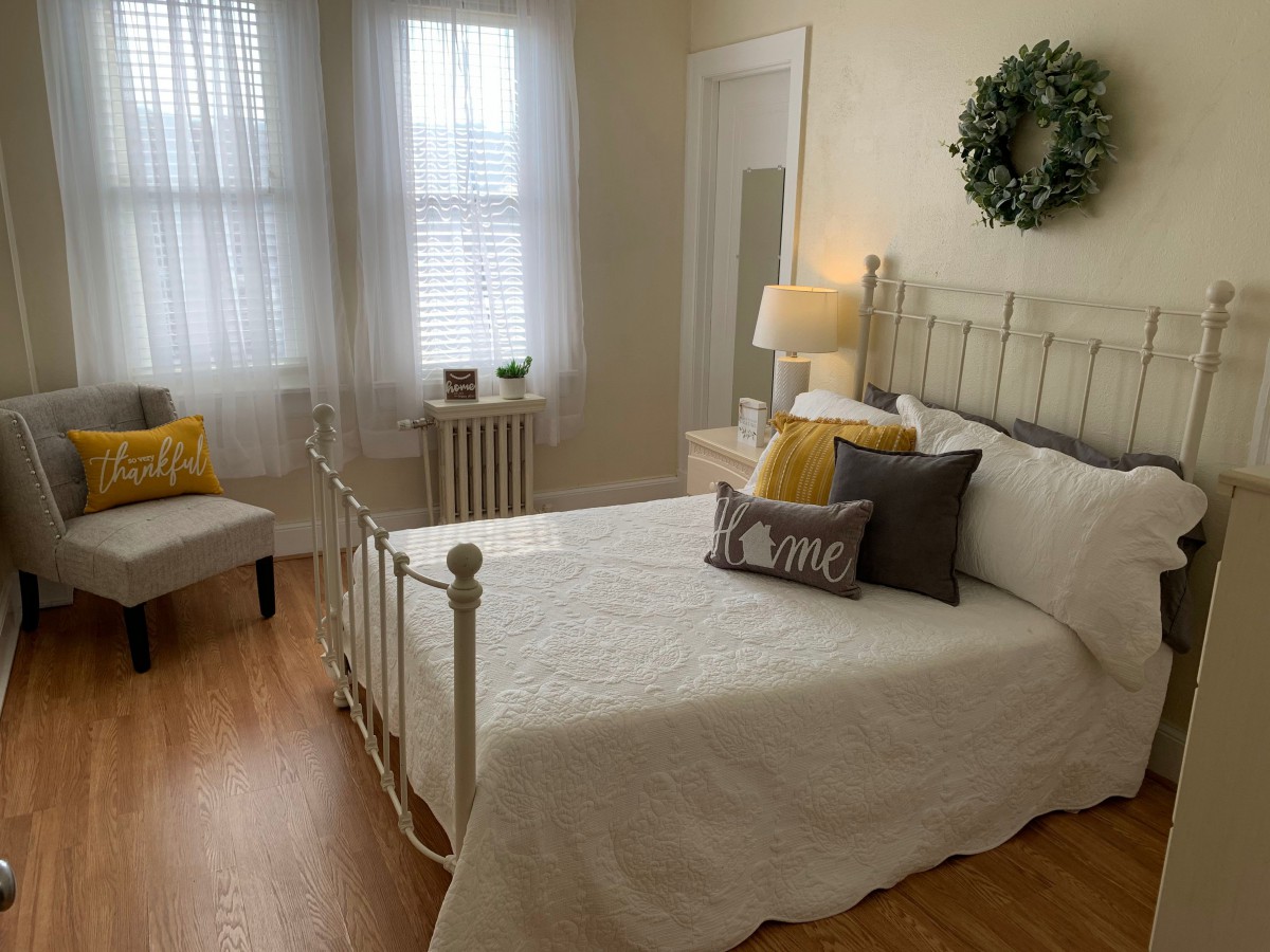 A cozy bedroom featuring a metal bedframe with white bedding, accented by yellow and gray cushions. Natural light filters through the windows, complemented by a bedside lamp. A decorative wreath hangs above the bed.