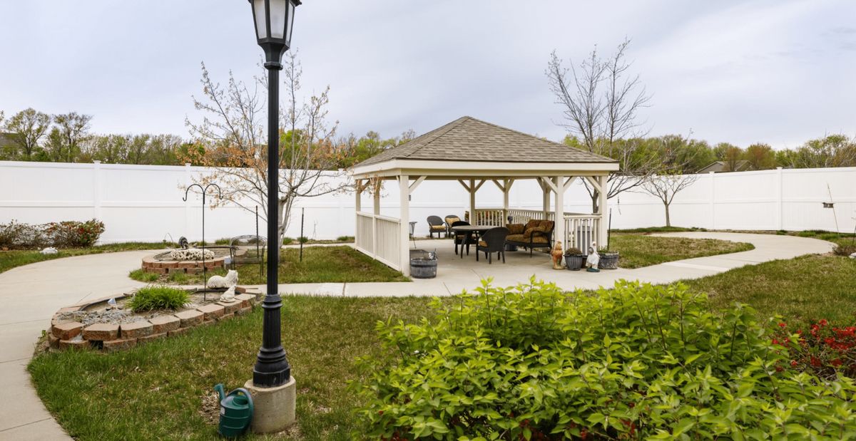 A backyard scene featuring a gazebo with patio furniture, surrounded by a well-maintained lawn and a white fence.