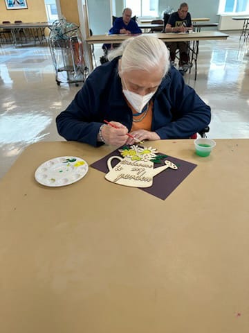 An elderly person engaged in a painting activity while wearing a mask, surrounded by art supplies.