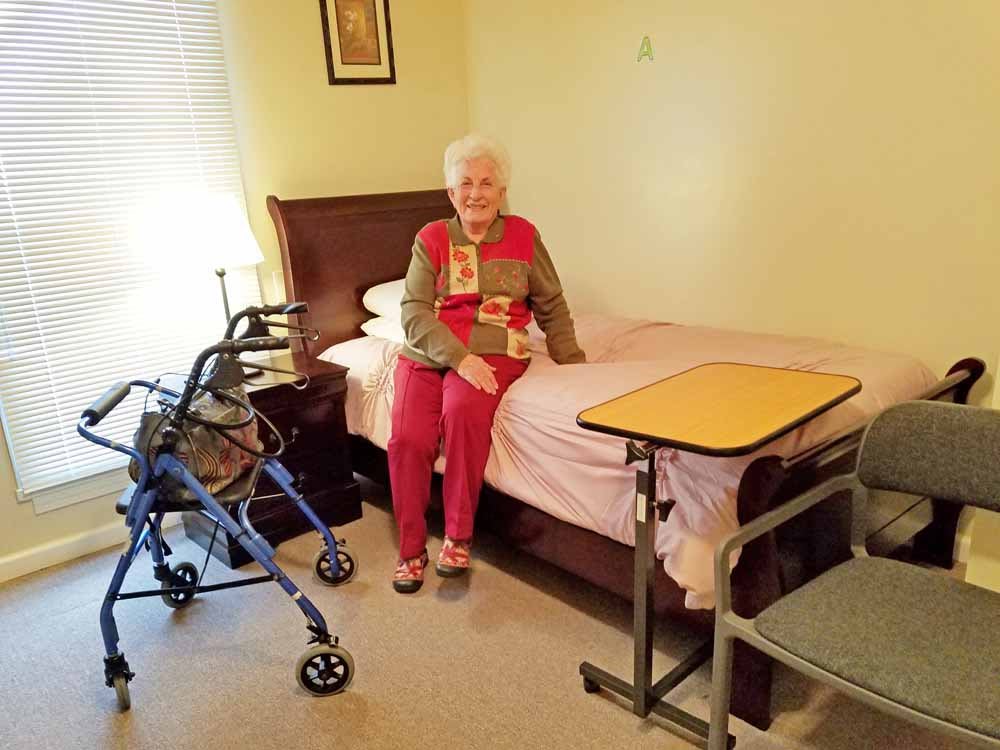 An elderly woman sitting on a bed in a care facility room with a walker nearby.