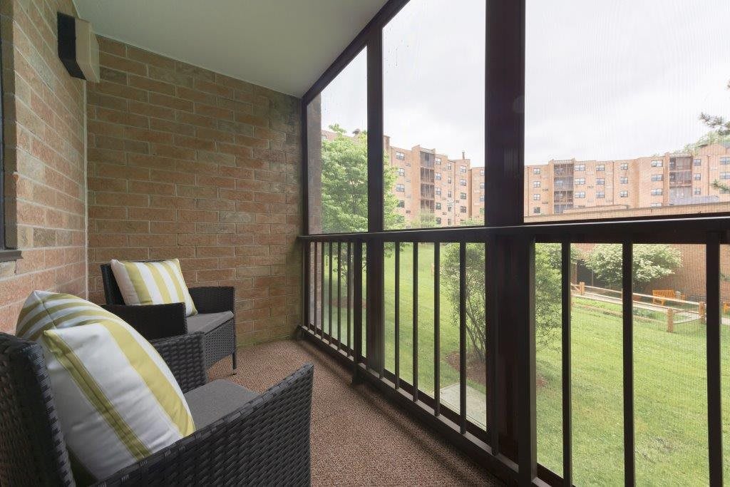A cozy screened balcony with wicker chairs and outdoor cushions, offering a view of nearby apartment buildings.