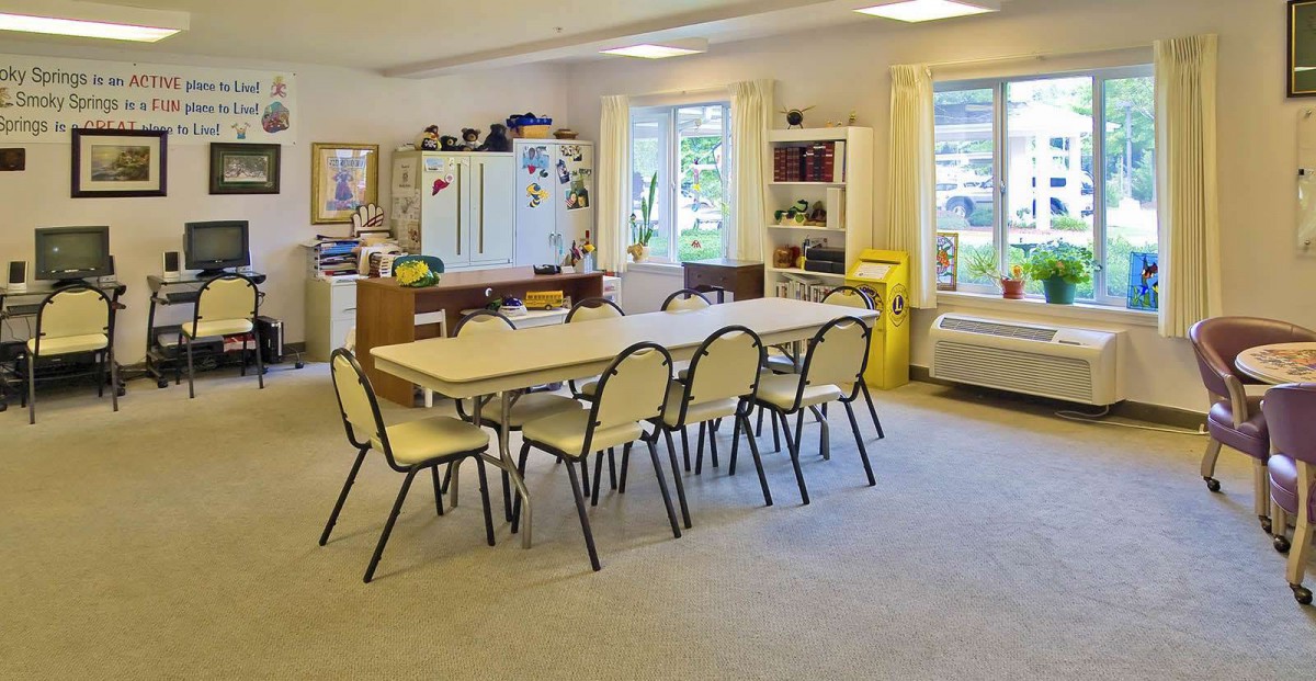 A classroom with tables and chairs arranged in the center, surrounded by shelves and windows.