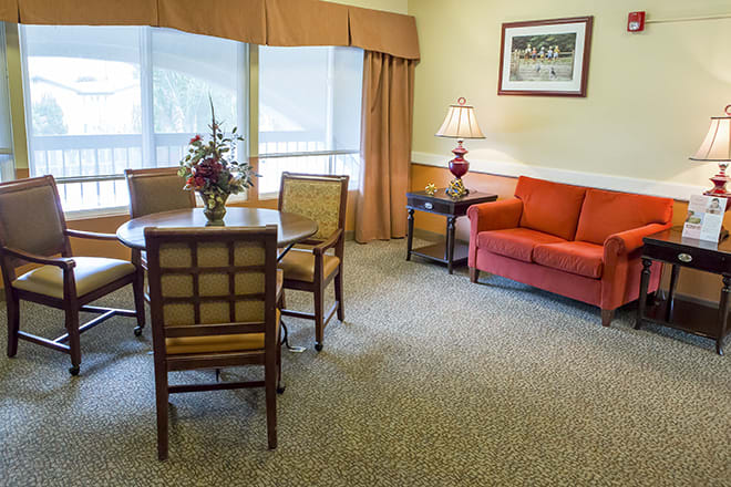 A cozy living room with a round table and chairs, an orange couch, a side table with a lamp, and large windows.