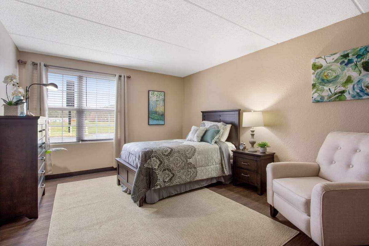 A cozy bedroom with a single bed, a nightstand with a lamp, and a comfortable chair by the window.