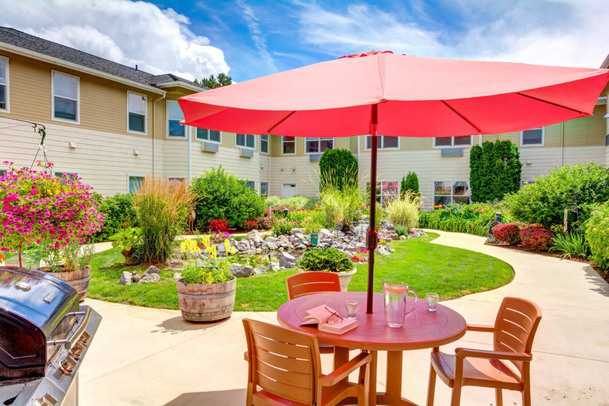 A sunny outdoor patio area with a red umbrella and wooden furniture surrounded by a lush garden.