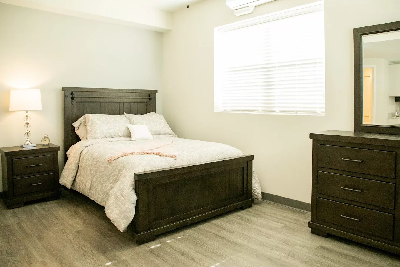 A minimalist bedroom with a wooden bed and dresser, featuring light walls and a large window.