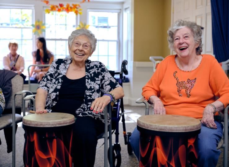 Two seniors enjoy a drumming activity indoors, both smiling and engaged.