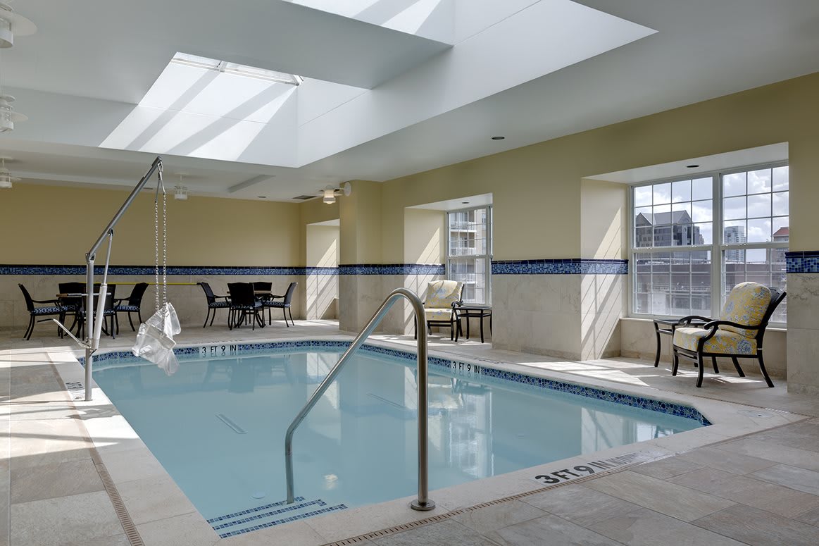 A serene indoor pool area with ample natural light from skylights and windows, surrounded by seating and tables.