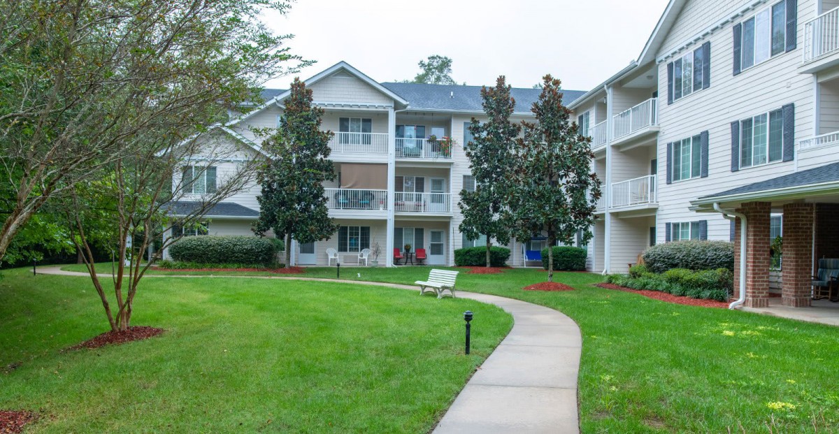 A residential apartment building with a well-maintained lawn and pathway surrounded by trees.