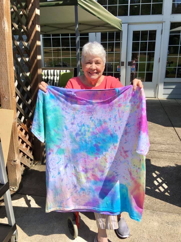 A senior woman proudly displays a colorful tie-dye t-shirt outdoors.