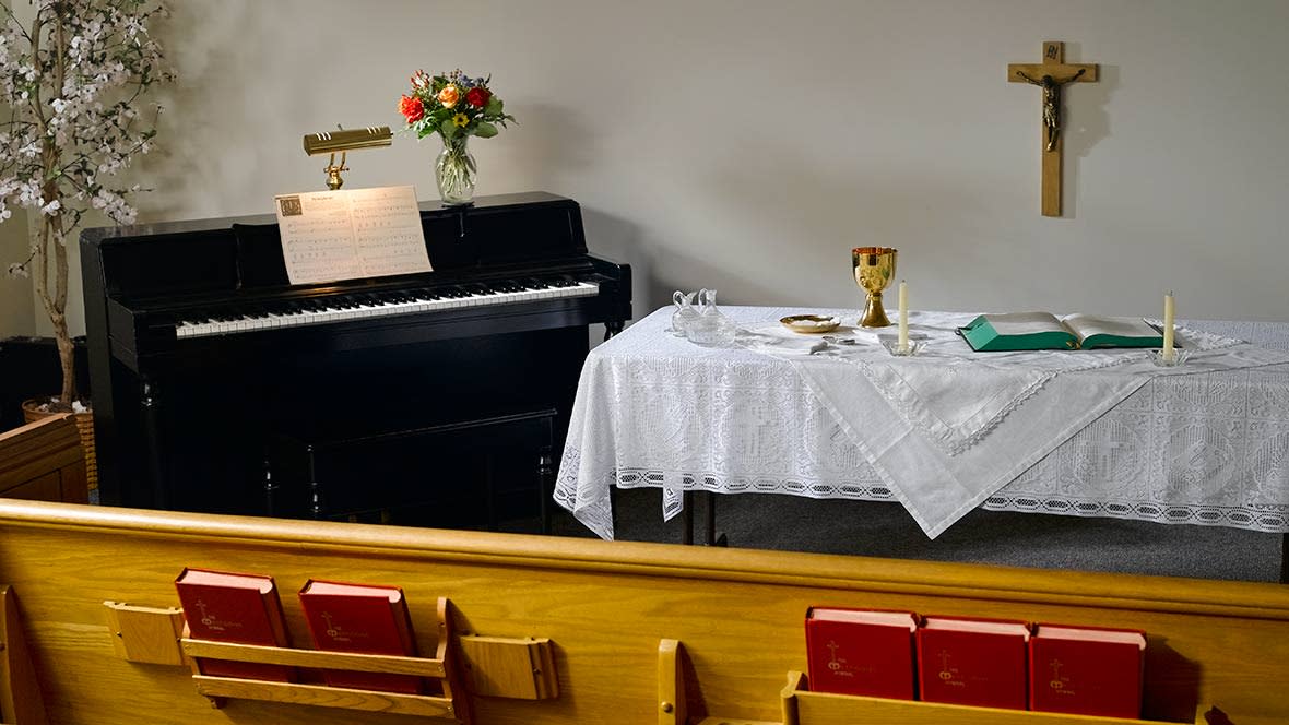 A church setting with a piano, flowers, and an altar table covered with a white cloth.