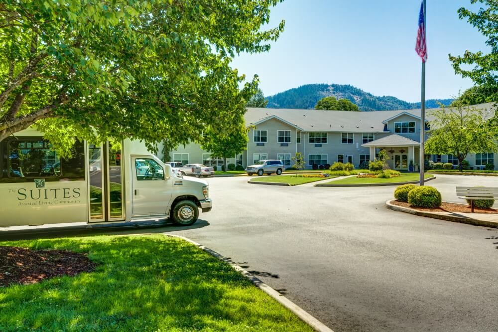A sunny day with a white vehicle parked near a building surrounded by greenery and mountains.