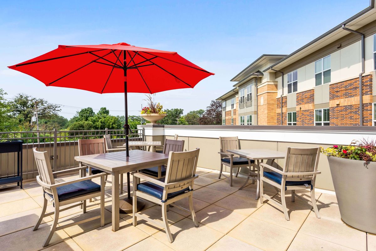 A deck with outdoor seating and a red umbrella near a building surrounded by trees.