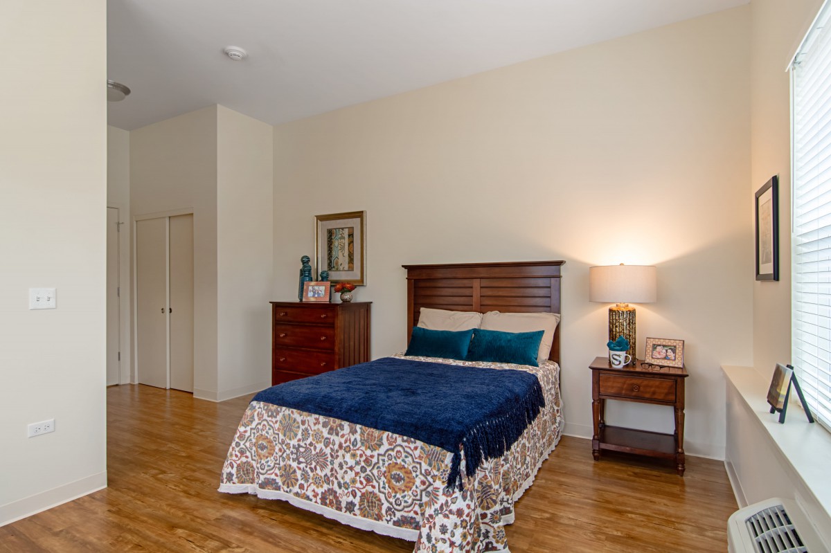 A cozy bedroom with a neatly made bed, wooden dresser, and a lamp on the nightstand.