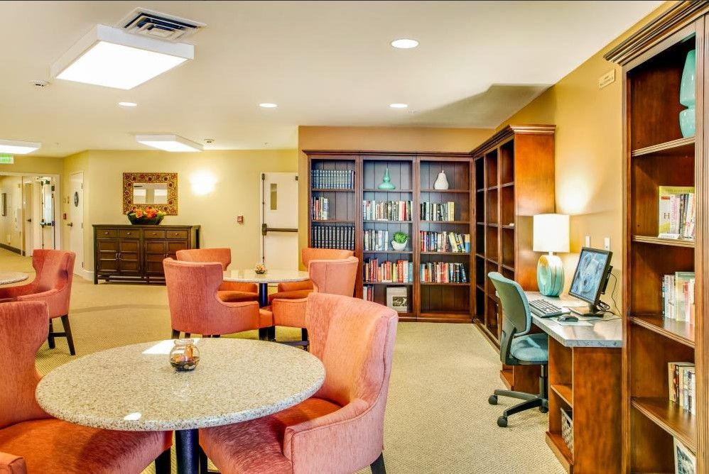 A cozy library or office space with bookshelves, a computer desk, round tables, and warm lighting.