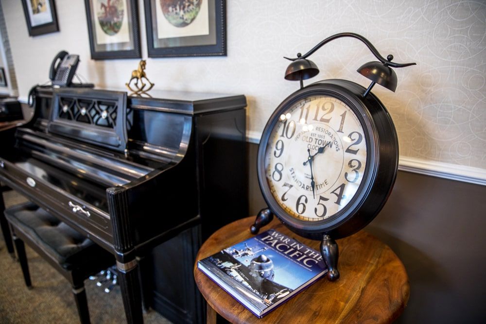A cozy interior with a vintage clock on a table beside a piano, accompanied by artwork and a magazine.