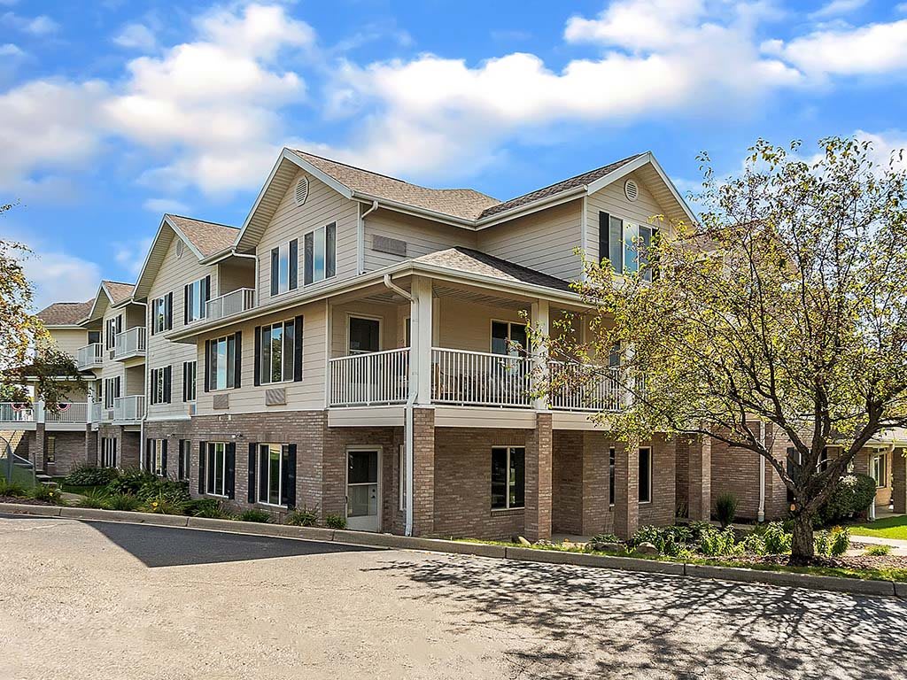 A two-story residential building with balconies and a tree-lined driveway under a clear blue sky.