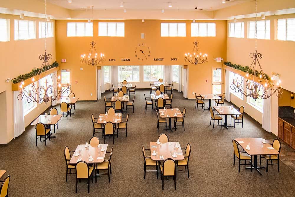 An elegant dining room with neatly arranged tables and chairs, lit by chandeliers and natural light from large windows.