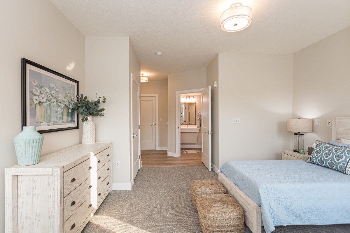 A minimalist bedroom featuring neutral tones, with a bed, dressers, and soft lighting.