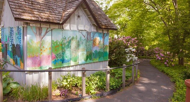 A quaint cabin with a colorful mural sits beside a lush garden and pathway under vibrant green trees.