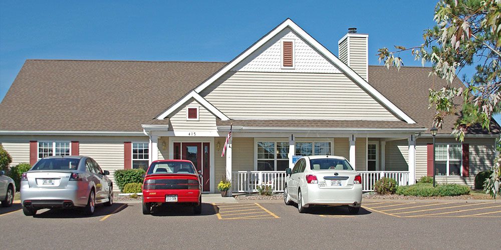 A small house with a front porch and two cars parked in the driveway on a sunny day.