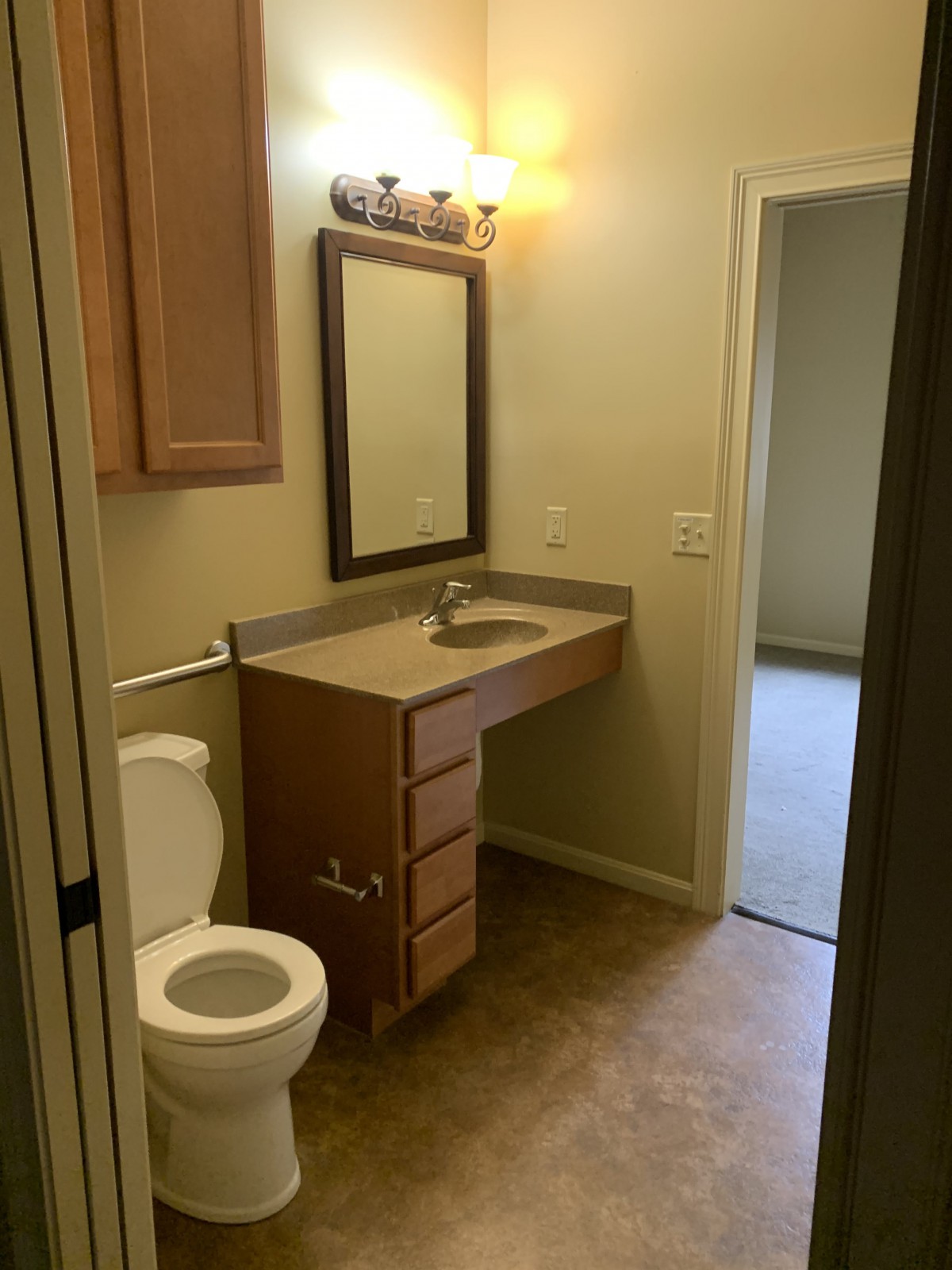 A small bathroom with a toilet, sink, and mirror, featuring wooden cabinets and a light fixture.